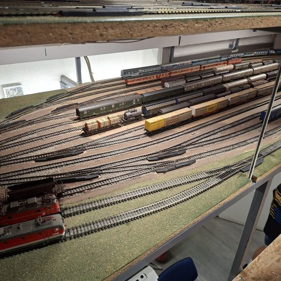 Ein Modelleisenbahn-Layout mit mehreren Gleisen und Zügen, einschließlich eines roten und weißen Zuges auf der linken Seite und verschiedener anderer Züge auf der rechten Seite.