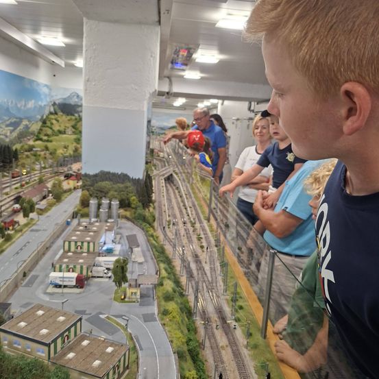 Eine Gruppe von Menschen, einschließlich Kindern, beobachtet eine Miniatur-Zugausstellung. Die Szene zeigt eine detaillierte Modellstadt mit Gebäuden, Straßen und einem Eisenbahnsystem.