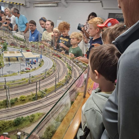 Eine Gruppe von Kindern und Erwachsenen versammelt sich um eine große Modelleisenbahnanlage und beobachtet die Miniaturzüge und die Landschaft. Die Anlage verfügt über mehrere Gleise, einen Bahnhof und verschiedene Gebäude.