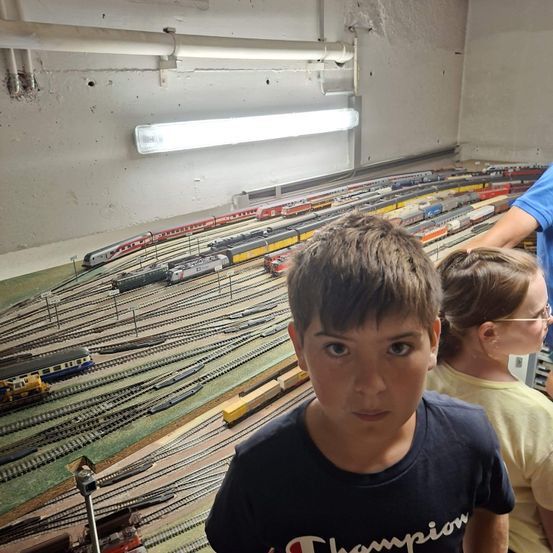 Ein Junge in einem Champion-Shirt steht vor einer Modelleisenbahn mit mehreren Gleisen und Zügen. Hinter ihm schaut ein Mädchen mit Brille auf die Züge.