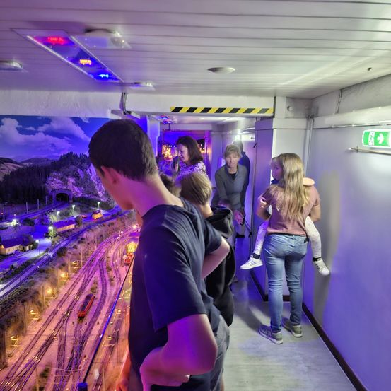 Eine Gruppe von Menschen steht in einem Tunnel mit einer Modelleisenbahnstrecke darunter. Der Tunnel hat ein lila Licht und eine Wandmalerei.