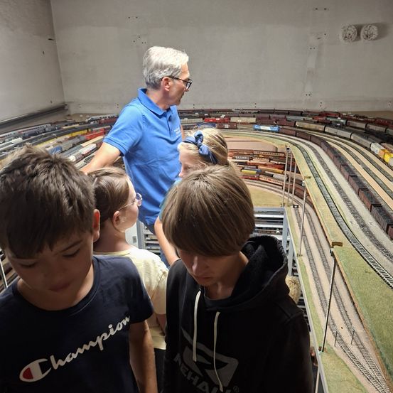 Eine Gruppe von Kindern und einem erwachsenen Mann betrachten eine Modelleisenbahn. Der Erwachsene trägt eine Brille und ein blaues Poloshirt. Die Kinder tragen Freizeitkleidung.