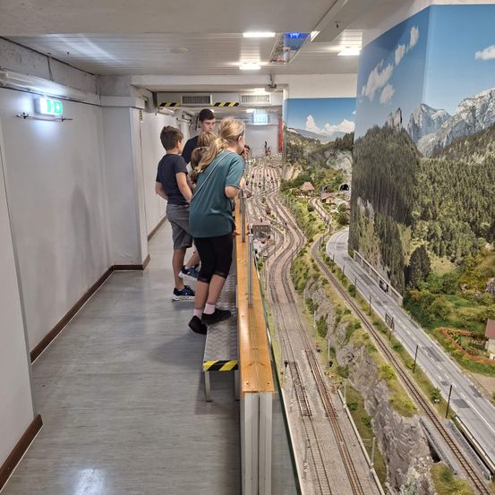 Eine Gruppe von Menschen steht auf einem Balkon und blickt auf eine Modelleisenbahn mit einer malerischen Kulisse von Bergen und einer Straße.