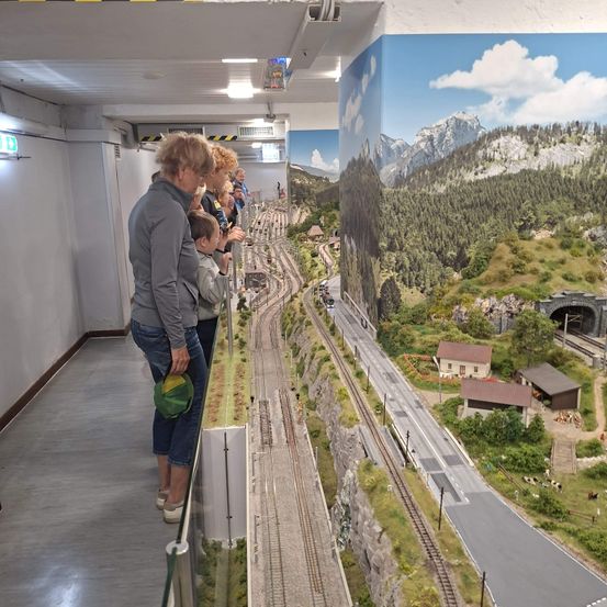 Eine Gruppe von Menschen, darunter Erwachsene und Kinder, beobachten ein Miniatur-Modelleisenbahn-Layout mit einer malerischen Kulisse von Bergen und einem kleinen Dorf.