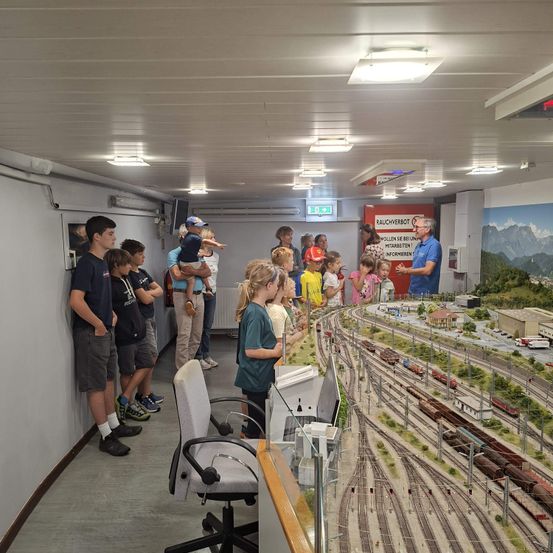 Eine Gruppe von Kindern und Erwachsenen versammelt sich um eine große Modelleisenbahn, wobei ein Erwachsener ihnen etwas erklärt. Die Modelleisenbahn steht auf einem Tisch, und es stehen Leute darum.