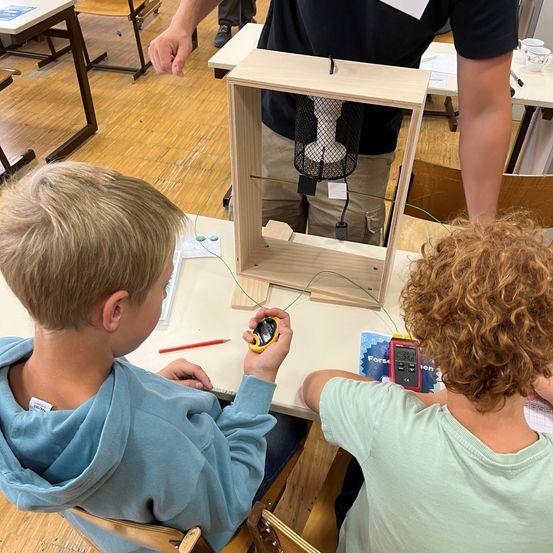 Zwei Kinder sitzen an einem Schreibtisch, eines benutzt ein Multimeter und das andere hält ein kleines elektronisches Gerät. Eine Holzkiste mit einem Draht und einem schwarzen Netz steht auf dem Tisch. Eine Person steht hinter ihnen.