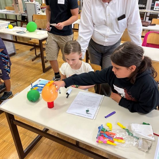 Zwei junge Mädchen sind in einem Klassenzimmer mit einem Wissenschaftsexperiment beschäftigt, wobei eines ein orangefarbenes Luftballon hält und das andere ein Papier. Ein Mann steht hinter ihnen und beobachtet.