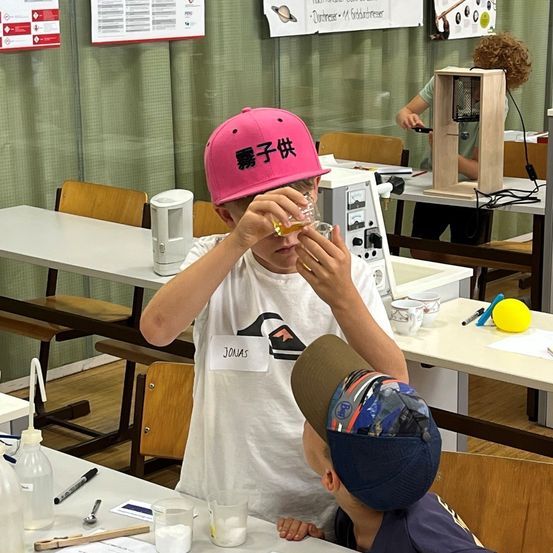 Ein junger Junge mit einer pinken Kappe untersucht eine Flüssigkeit in einem kleinen Glas, während ein anderer Junge mit einer blauen Kappe zusieht. Sie sitzen an einem Tisch in einem Klassenzimmer mit verschiedenen Lehrmaterialien und Geräten um sie herum.