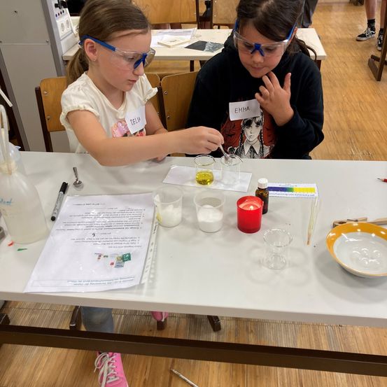 Zwei junge Mädchen in Schutzbrillen führen ein wissenschaftliches Experiment an einem Tisch mit verschiedenen Laborgeräten durch. Eines der Mädchen trägt den Namen 'Delia' und das andere 'Emma'.