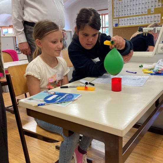 Zwei junge Mädchen sitzen an einem Tisch, eine hält einen grünen Ballon und die andere beobachtet. Sie sind von verschiedenen Bastelmaterialien umgeben. Ein Mann steht hinter ihnen und beobachtet.
