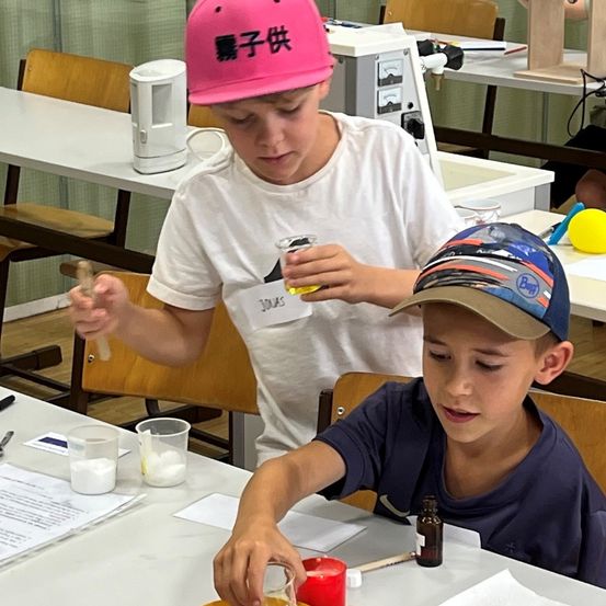 Zwei Jungen führen ein wissenschaftliches Experiment in einem Klassenzimmer durch. Ein Junge hält eine Pipette und ein Reagenzglas, während der andere Junge ein kleines Glas und eine Kerze hält.