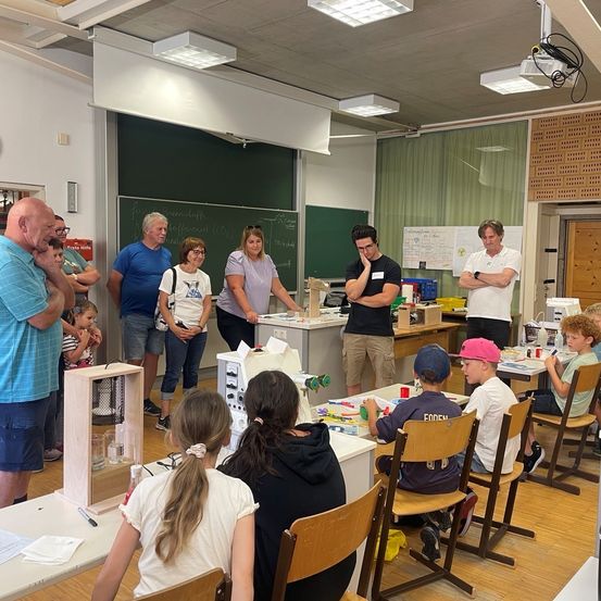 Eine Gruppe von Erwachsenen und Kindern in einem Klassenzimmer, wobei einige Personen stehen und andere um Tische herum sitzen und eine Aktivität mit einer Maschine durchführen.