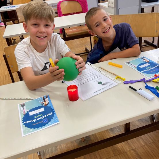 Zwei Jungen sitzen an einem Tisch in einem Klassenzimmer, einer hält einen grünen Ballon und der andere lächelt. Der Tisch hat Papiere, eine Kerze und andere Gegenstände.