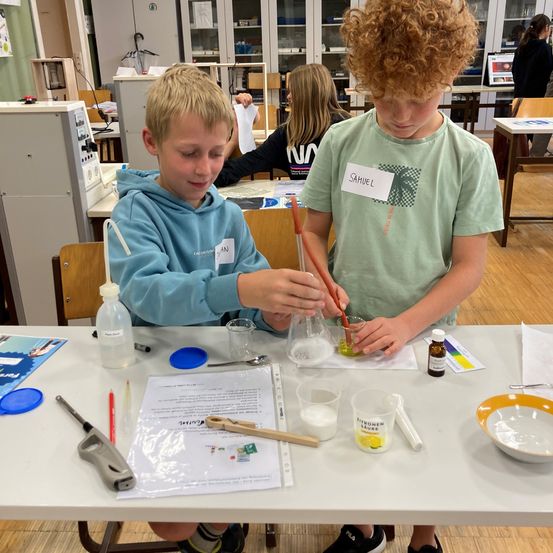 Zwei junge Jungen führen ein Chemieexperiment in einem Klassenzimmer durch. Ein Junge gießt eine Flüssigkeit in ein Becherglas, während der andere eine Pipette hält. Verschiedene Laborgeräte und Materialien liegen auf dem Tisch.