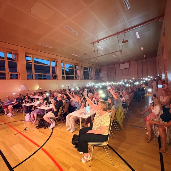 Eine Gruppe von Menschen in einer Halle, einige halten ihre Telefone mit eingeschalteten Lichtern hoch, möglicherweise machen sie ein Gruppenfoto.