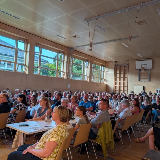 Eine große Gruppe von Menschen sitzt in einem Raum mit großen Fenstern, Tischen und Stühlen. Einige Leute schauen auf eine Präsentation an einer Leinwand.
