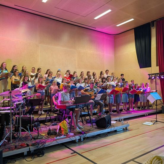 Ein Chor junger Menschen, Jungen und Mädchen, tritt auf einer Bühne mit Musikinstrumenten und pinker Beleuchtung auf.
