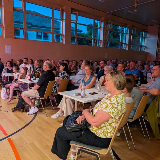 Eine große Gruppe von Menschen sitzt in einem Saal, einige stehen, und alle schauen auf eine Bühne. Der Saal hat große Fenster und einen Holzboden.