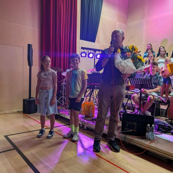 Ein Mann steht mit einem Blumenstrauß auf der Bühne und spricht eine Gruppe von Kindern in einem Schulauditorium an. Hinter ihm spielt eine Band, und eine Menge Schüler schaut zu.