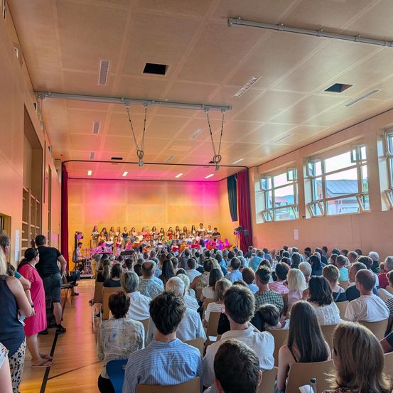 Ein großer Innensaal mit einem Chor auf der Bühne, beleuchtet von rosa und gelben Lichtern, mit einer Menge Menschen, die vor ihnen in Stühlen sitzen.