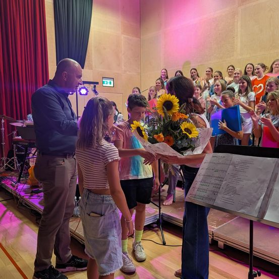 Eine Gruppe von Kindern und Erwachsenen versammelt sich in einem Raum, wobei ein Mann einer jungen Mädchen Blumen überreicht. Hinter ihnen singt ein Chor, und auf der Bühne sind Musikinstrumente aufgebaut.
