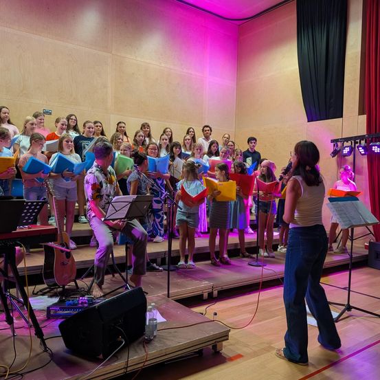 Ein Chor junger Menschen steht mit Musikinstrumenten und bunten Ordnern auf der Bühne und singt unter rosa Lichtern.