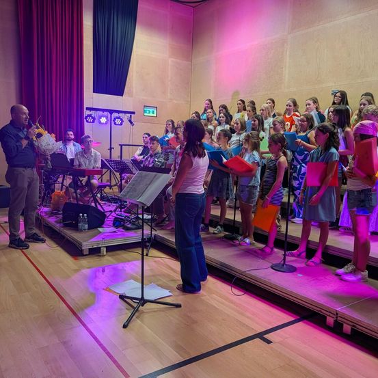 Ein Chor aus Kindern und Erwachsenen tritt mit einem Dirigenten und Musikern auf der Bühne auf, beleuchtet von lila Lichtern.