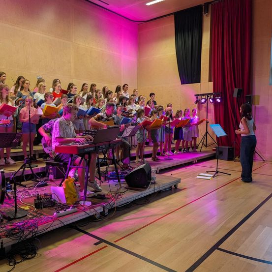Ein Chor junger Menschen tritt mit Musikinstrumenten und einem Dirigenten auf der Bühne auf. Die Bühne ist mit rosa Lichtern beleuchtet.