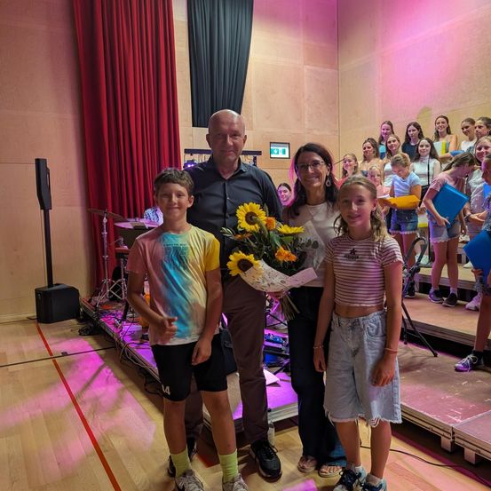 Ein Mann und zwei Kinder stehen mit Blumen auf der Bühne und lächeln für ein Foto. Dahinter sitzen eine Gruppe von Kindern und Erwachsenen im Publikum.