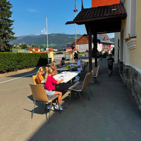 Eine Gruppe von Kindern sitzt an Tischen draußen und macht Kunstarbeiten. Die Tische sind unter einem Vordach aufgestellt. Im Hintergrund befindet sich ein Gebäude, eine Straße und eine Aussicht auf Berge.