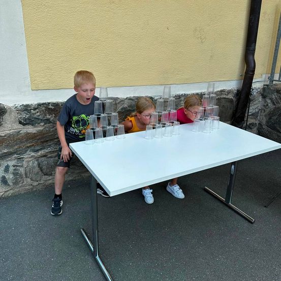 Drei Kinder spielen ein Spiel mit Bechern auf einem weißen Tisch und lehnen sich vor, um sie auszubalancieren.
