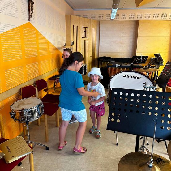 Zwei Mädchen sind in einem Musikraum, eine lehrt die andere, wie man Schlagzeug spielt. Verschiedene Musikinstrumente sind um sie herum.