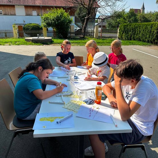 Eine Gruppe von Kindern sitzt draußen um einen Tisch herum und macht etwas Zeichnen und Schreiben. Sie haben Marker, Papiere und eine Flasche Saft auf dem Tisch. Hinter ihnen steht ein Gebäude mit einem Zaun und einigen Bäumen.