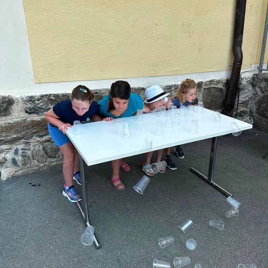 Vier Kinder spielen ein Spiel mit Bechern auf einem weißen Tisch, mit Bechern, die um sie herum auf dem Boden verstreut sind.
