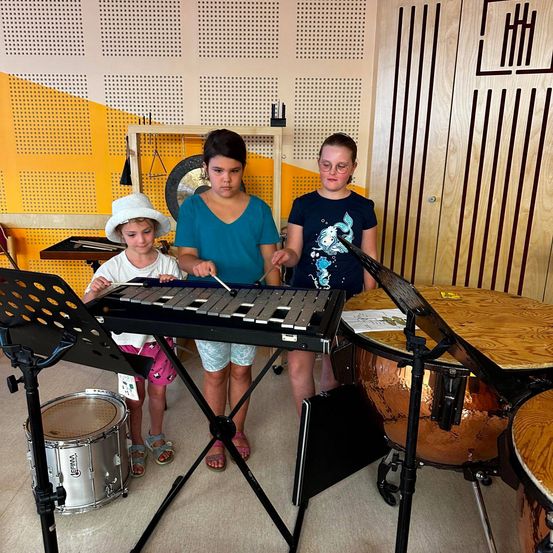Drei Kinder in einem Musikraum, eines spielt Xylophon, ein anderes hält einen Drumstick und das dritte beobachtet. Verschiedene Musikinstrumente sind um sie herum angeordnet.
