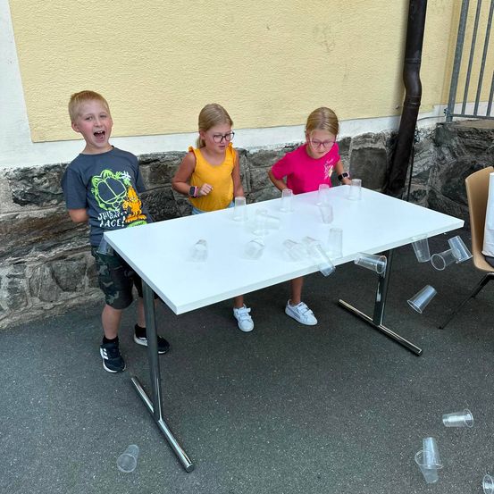 Drei Kinder stehen um einen Tisch herum, auf dem sich Becher befinden, von denen einige auf den Boden gefallen sind. Der Junge lacht, während die beiden Mädchen die Becher betrachten.