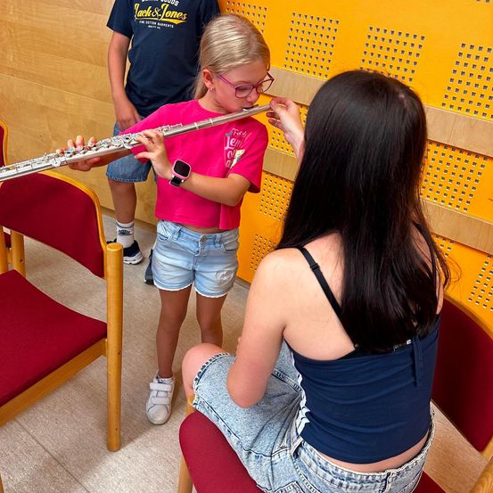 Ein junges Mädchen in einem rosa T-Shirt spielt Flöte, während eine Frau in einem blauen Oberteil ihr hilft, es anzupassen. Dahinter steht ein Junge in einem blauen T-Shirt und beobachtet.