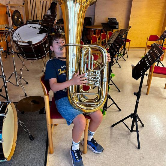 Ein junger Junge spielt eine große Messingtuba in einem Musikraum, umgeben von verschiedenen Instrumenten und Stühlen.