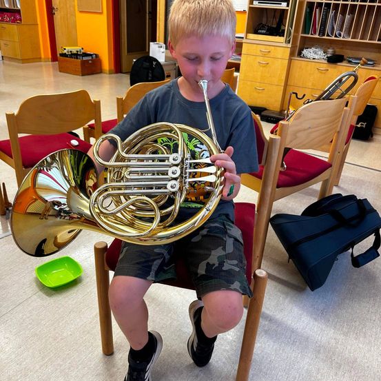 Ein junger Junge sitzt auf einem Stuhl in einem Musikzimmer, hält ein Waldhorn und spielt es. Er trägt ein blaues T-Shirt und Shorts.