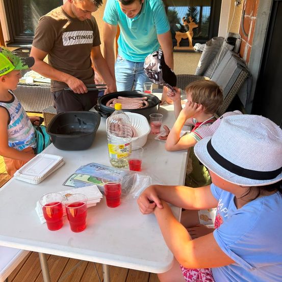 Eine Familie bereitet Essen auf einem Tisch im Freien zu. Ein Mann grillt Würste, während ein anderer Mann und ein Kind zusehen. Der Tisch hat Getränke, Essen und Besteck.