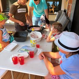 Eine Familie bereitet Essen auf einem Tisch im Freien zu. Ein Mann grillt Würste, während ein anderer Mann und ein Kind zusehen. Der Tisch hat Getränke, Essen und Besteck.
