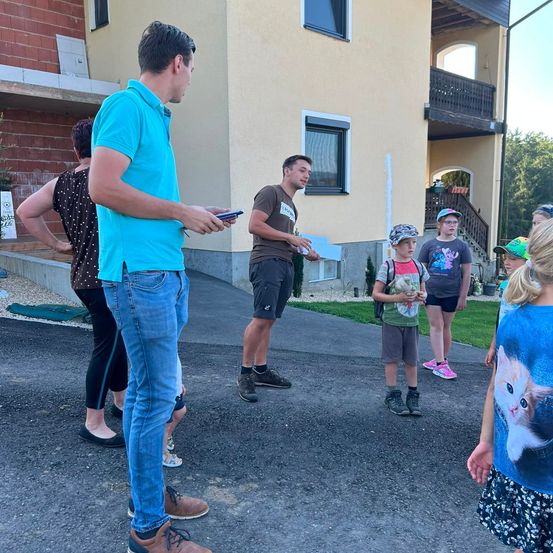 Eine Gruppe von Menschen, einschließlich Erwachsener und Kindern, steht vor einem Gebäude mit Balkon. Ein Mann hält ein Telefon und ein anderer ein Stück Papier. Die Kinder tragen Mützen und Turnschuhe.