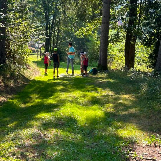 Eine Gruppe von Menschen, einschließlich Kindern, steht in einem Grasbereich mit Bäumen. Ein Mann in einem blauen Hemd spricht mit ihnen.