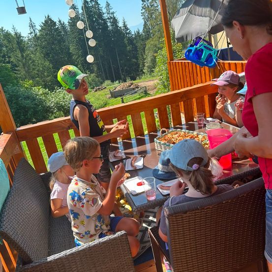 Eine Gruppe von Kindern und einer erwachsenen Frau sitzen auf einer Holzterrasse und genießen eine Mahlzeit. Die Kinder tragen Hüte und Brillen, und es gibt einen Tisch mit Essen, Getränken und einem Korb. Die Terrasse ist von Bäumen und Gras umgeben.