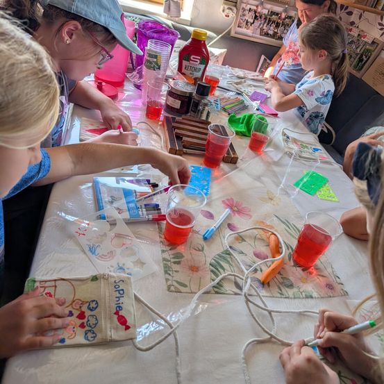 Eine Gruppe von Kindern sitzt um einen Tisch herum und ist in einer Bastelaktivität vertieft. Sie haben verschiedene Bastelutensilien verteilt, darunter Becher mit farbiger Flüssigkeit, Marker und kleine Beutel. Ein Kind hält einen Beutel mit dem Namen 'Sophie' darauf.