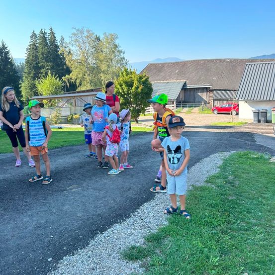 Eine Gruppe von Kindern und Erwachsenen steht auf einem Schotterweg in einer ländlichen Umgebung, mit Bäumen und einem Bauernhaus im Hintergrund.