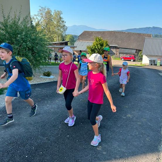Eine Gruppe von Kindern, einige mit Mützen und Rucksäcken, gehen eine Einfahrt hinunter, mit Häusern und Bergen im Hintergrund.