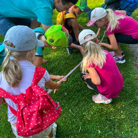Mehrere Kinder und Erwachsene sind auf einem Grasfeld versammelt, einige knien und andere stehen. Sie sind in einer Aktivität verstrickt, möglicherweise messen sie etwas mit einem Maßband.