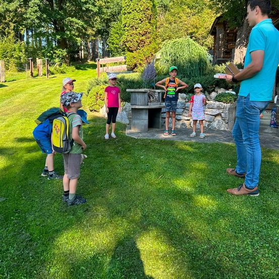 Eine Gruppe von Kindern und ein erwachsener Mann stehen auf einem Rasen, wobei der Erwachsene ein Buch hält und mit den Kindern spricht.