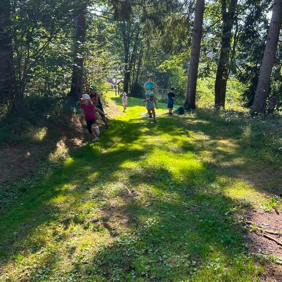 Eine Gruppe von Kindern und Erwachsenen läuft auf einem Grasweg im Wald. Einige tragen Mützen und Turnschuhe. Hohe Bäume umgeben das Gebiet.